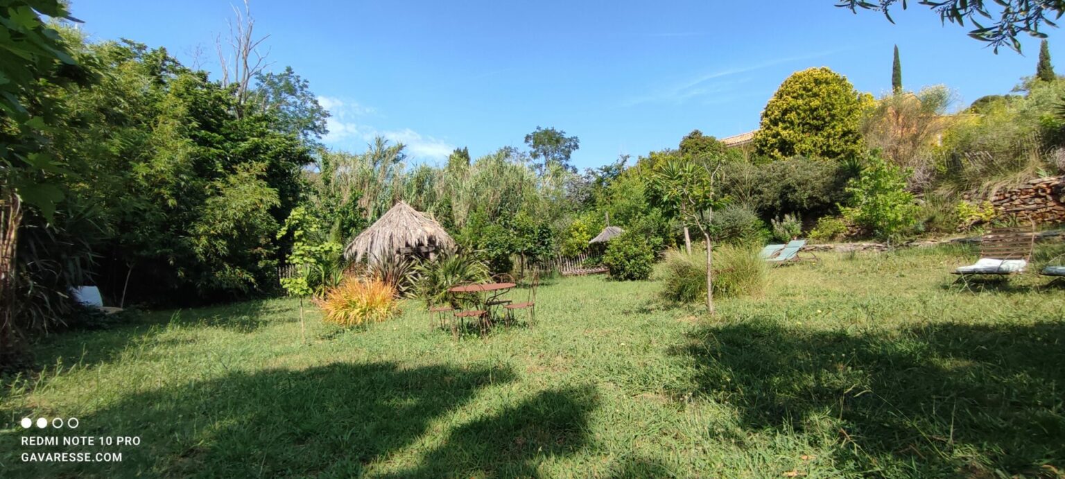 Vaste jardin paysagé méditerranéen de la Maison Gavaresse au Pradet, avec cabane pour enfants et mobilier extérieur, sous un ciel bleu.